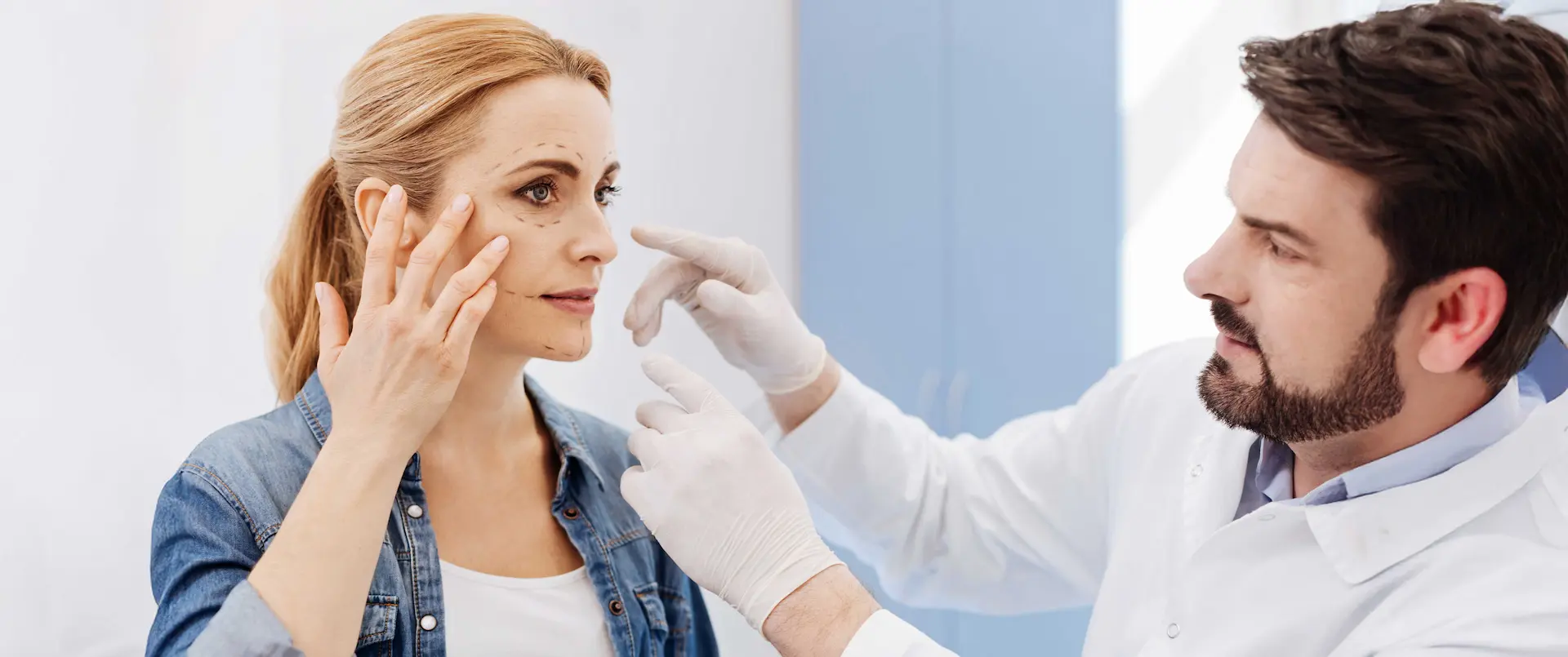 Medico chirurgo con guanti bianchi esamina una paziente con segni pre-operatori sul viso durante un consulto di chirurgia plastica nel centro medico